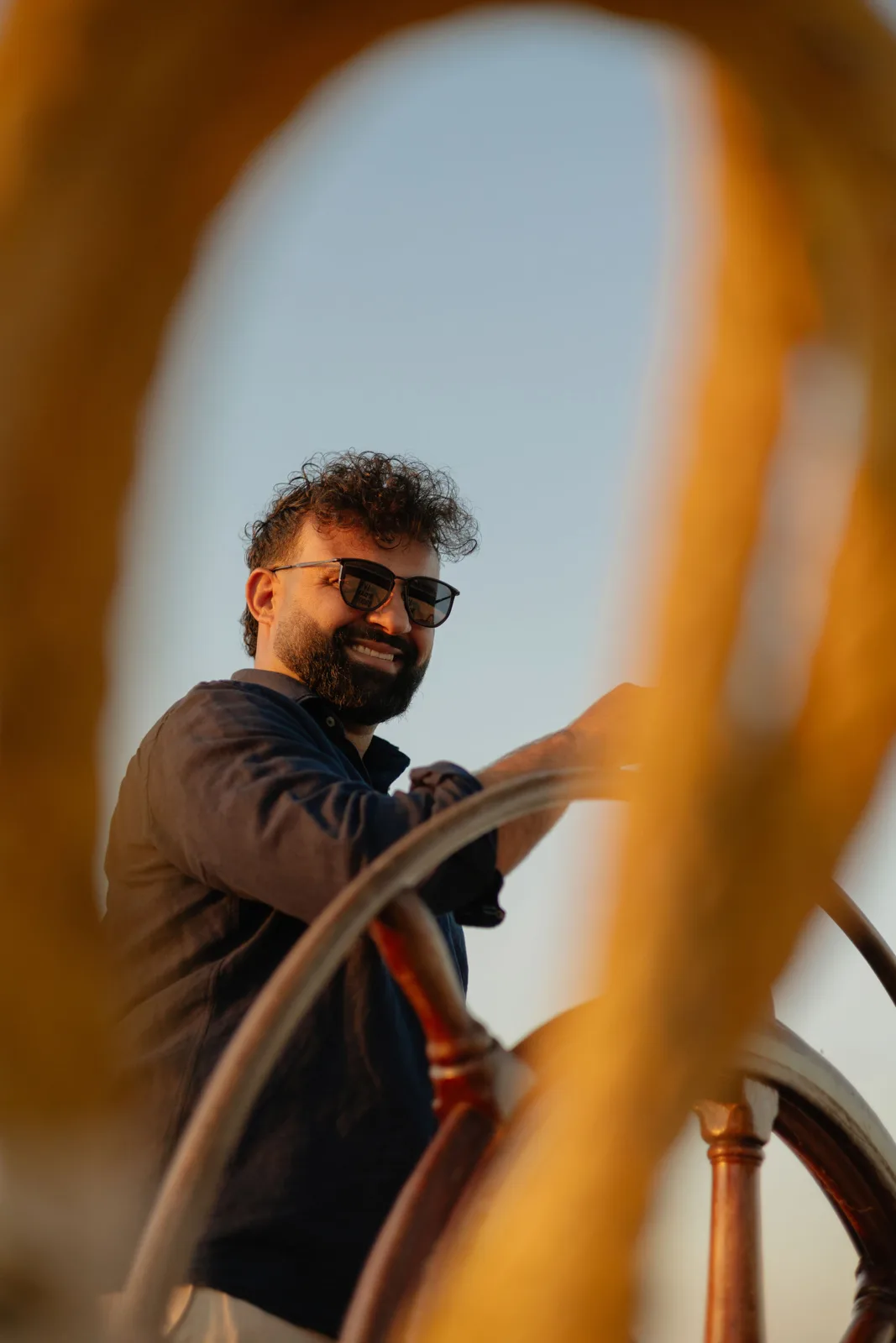 Amanj smiling at the helm of a ship, seen through the wheel